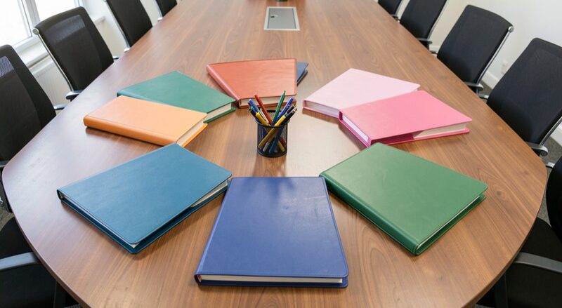 Six identical document folders in different colours arranged in a circle on a conference table, pens at the centre